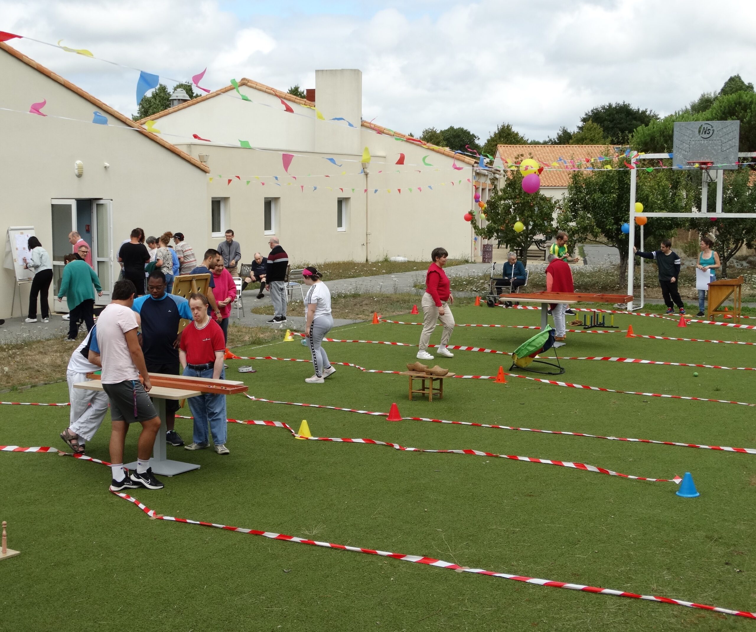 Les résidents du Centre d'Habitat d'Aizenay participent à des olympiades en extérieur. Plusieurs ateliers sont proposées. Des guirlandes de fanions décorent la cour.